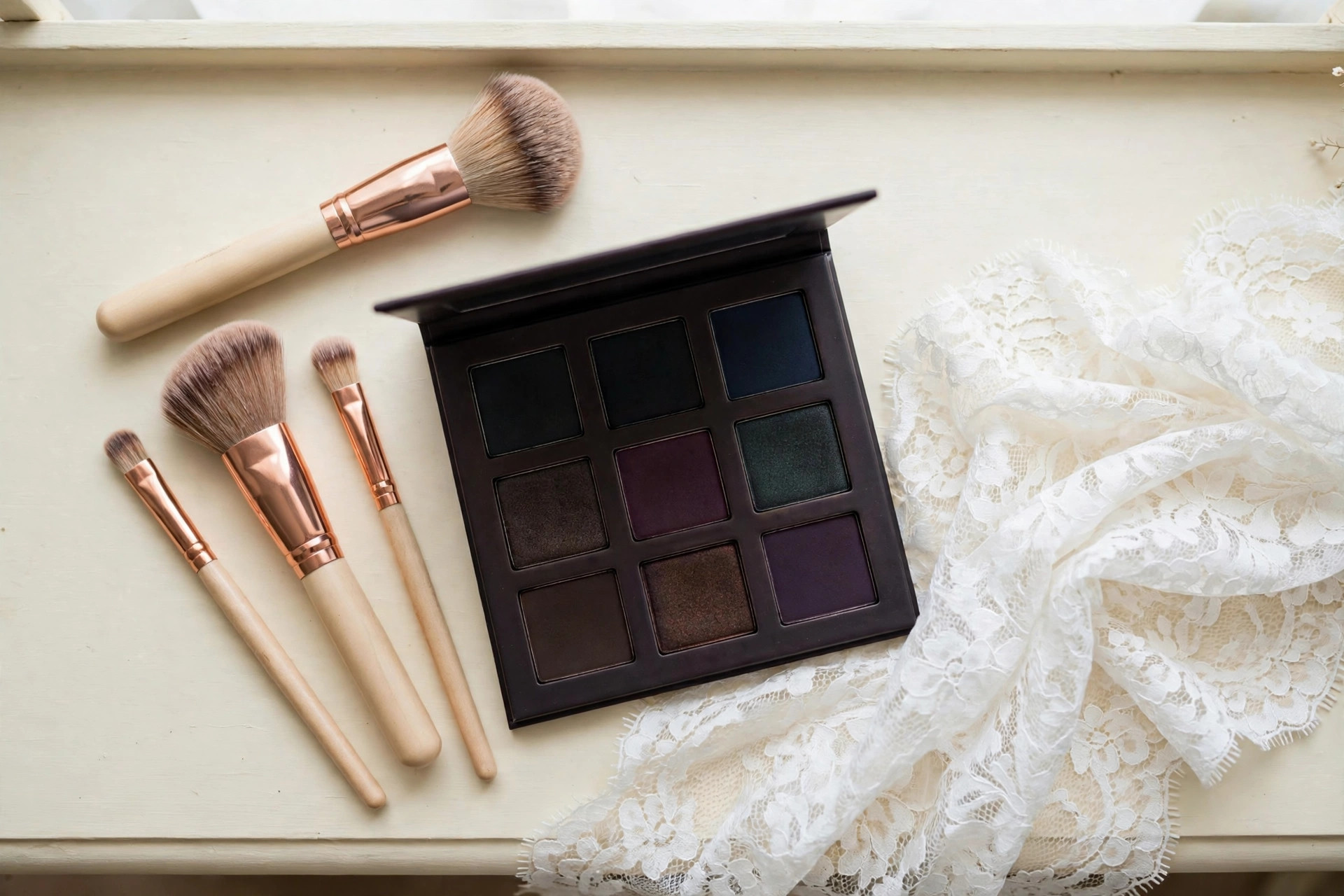 Flat-lay of makeup brushes, a dark eyeshadow palette and white lace fabric on a cream vanity table in soft daylight